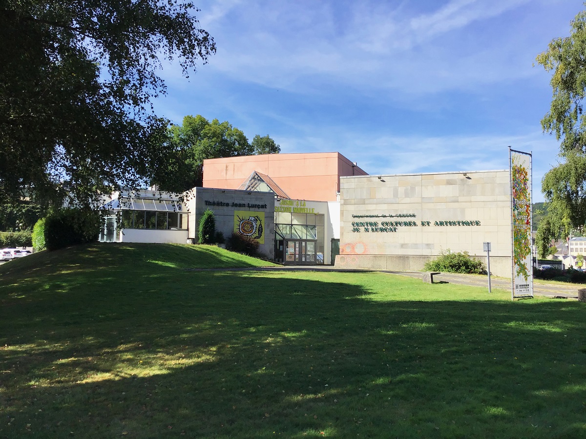 Au premier plan une pelouse verte où passe le chemin d’accès au théâtre. Au second plan, le bâtiment en pierre grise et verrières. Sur la partie droite est inscrit « CENTRE CULTUREL ET ARTISTIQUE JEAN LURÇAT ». Au centre l’entrée vitrée du théâtre à gauche de laquelle est inscrit "Théâtre Jean Lurçat" au-dessus d’une reproduction d’une œuvre picturale de Jean Lurçat. Surplombant l’entrée vitrée du théâtre on voit les murs de la salle en forme de cube rouge.